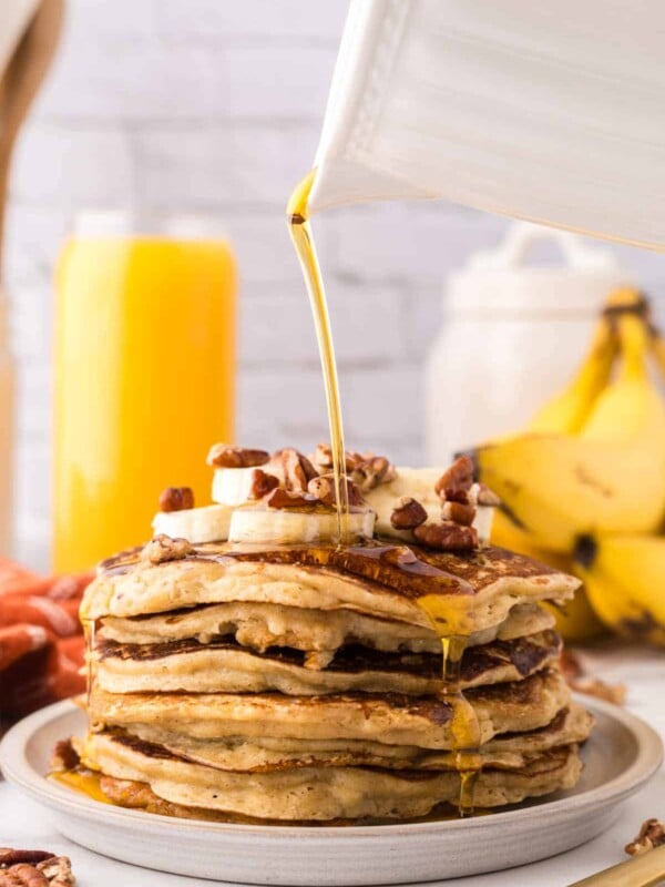 side view of a stack of banana pancakes on a round plate with walnuts and bananas with syrup being poured on top