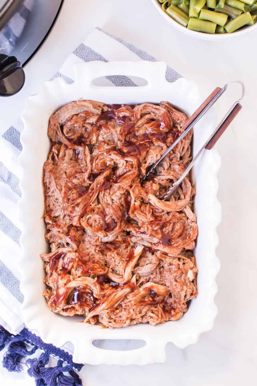 white casserole dish with barbecue pulled pork tongs and bowl of green beans.