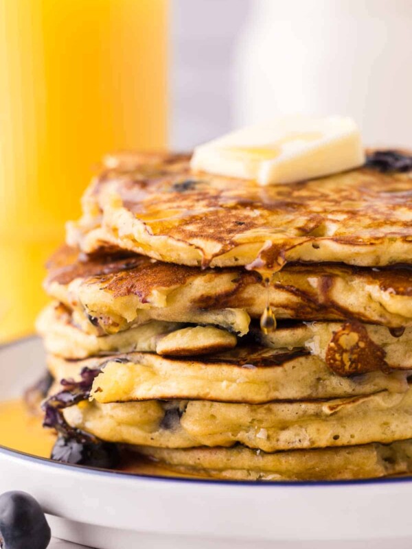 side view of a round white plate with a blue outer edge with a stack of fluffy blueberry pancakes with butter and syrup