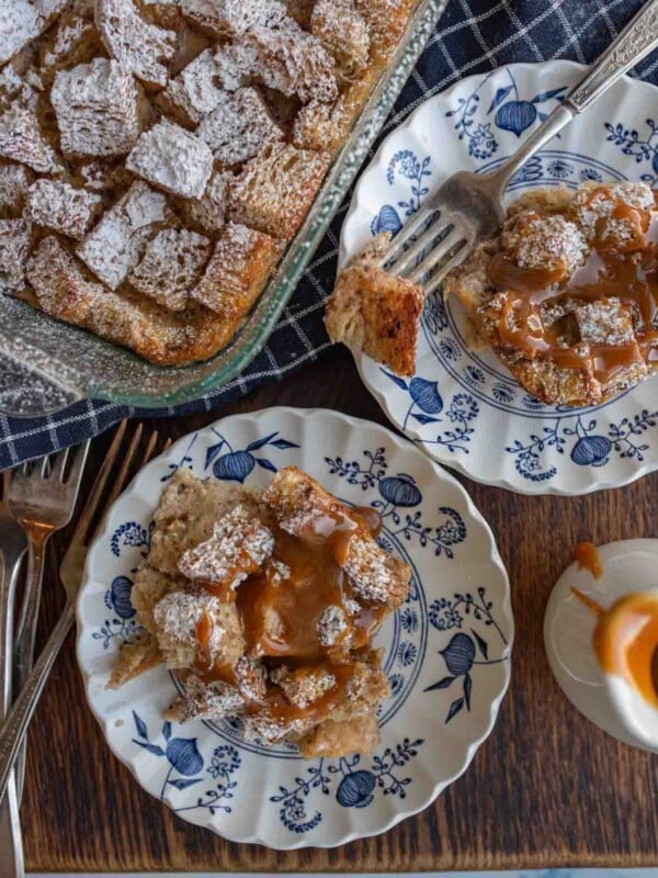 clear baking dish of squares of bread pudding with powdered sugar atop and served onto classic china dishes on the side with caramel on top