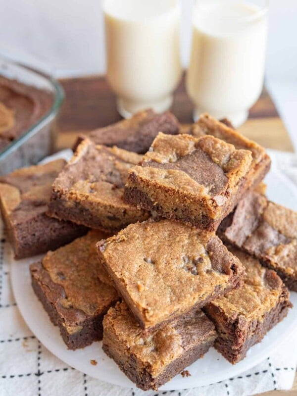 A plate stacked with homemade chocolate chip brownies, featuring a golden-brown top with visible chocolate chips reminiscent of cookies mixed with brownies. In the background, two glasses of milk and a dish of more decadent treats rest on a wooden surface.