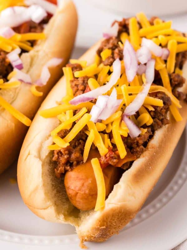 chili dogs recipe over round white plates with cheese and onions on top.