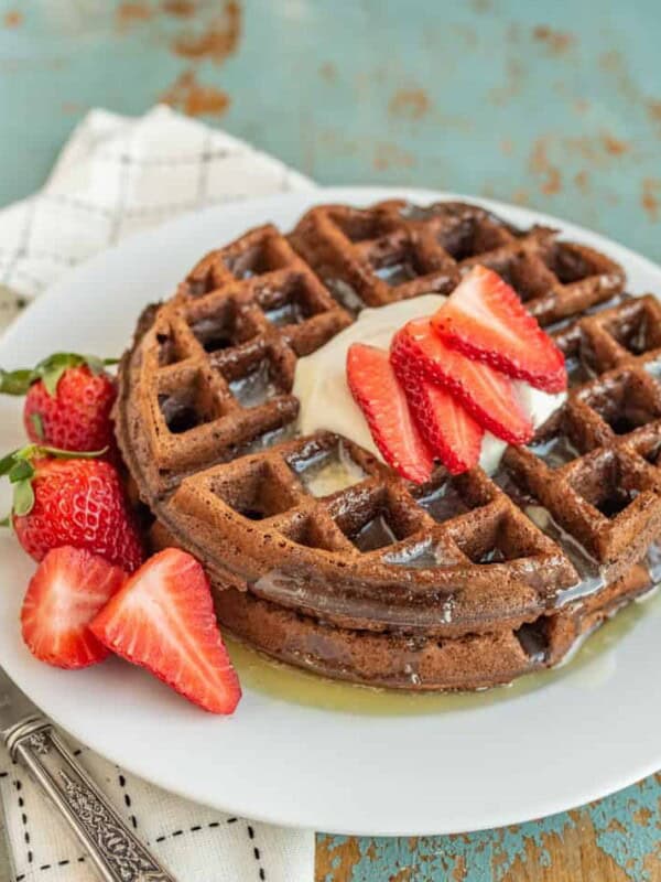 choclate waffle on a white plate with whole strawberries beside them and sliced strawberries, butter, and syrup on top.