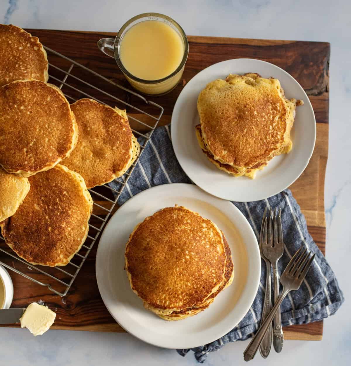 A wooden table with a stack of golden pancakes on a white plate, accompanied by butter and a glass of orange juice. More pancakes rest on a cooling rack. A blue napkin and silverware are placed nearby.