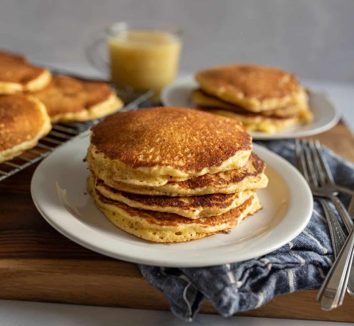 A stack of golden-brown pancakes on a white plate, accompanied by a blue cloth napkin and two forks. A second plate with pancakes and a glass mug with yellow liquid are in the background, all set on a wooden board.