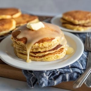 A stack of cornmeal pancakes topped with melting butter and syrup on a white plate. There are more pancakes in the background on a cooling rack and a separate plate. Forks are placed beside the dish on a striped cloth.