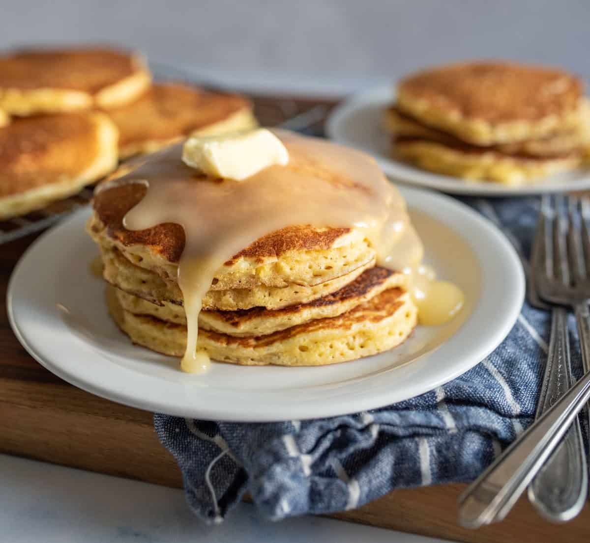 A stack of cornmeal pancakes topped with melting butter and syrup on a white plate. There are more pancakes in the background on a cooling rack and a separate plate. Forks are placed beside the dish on a striped cloth.