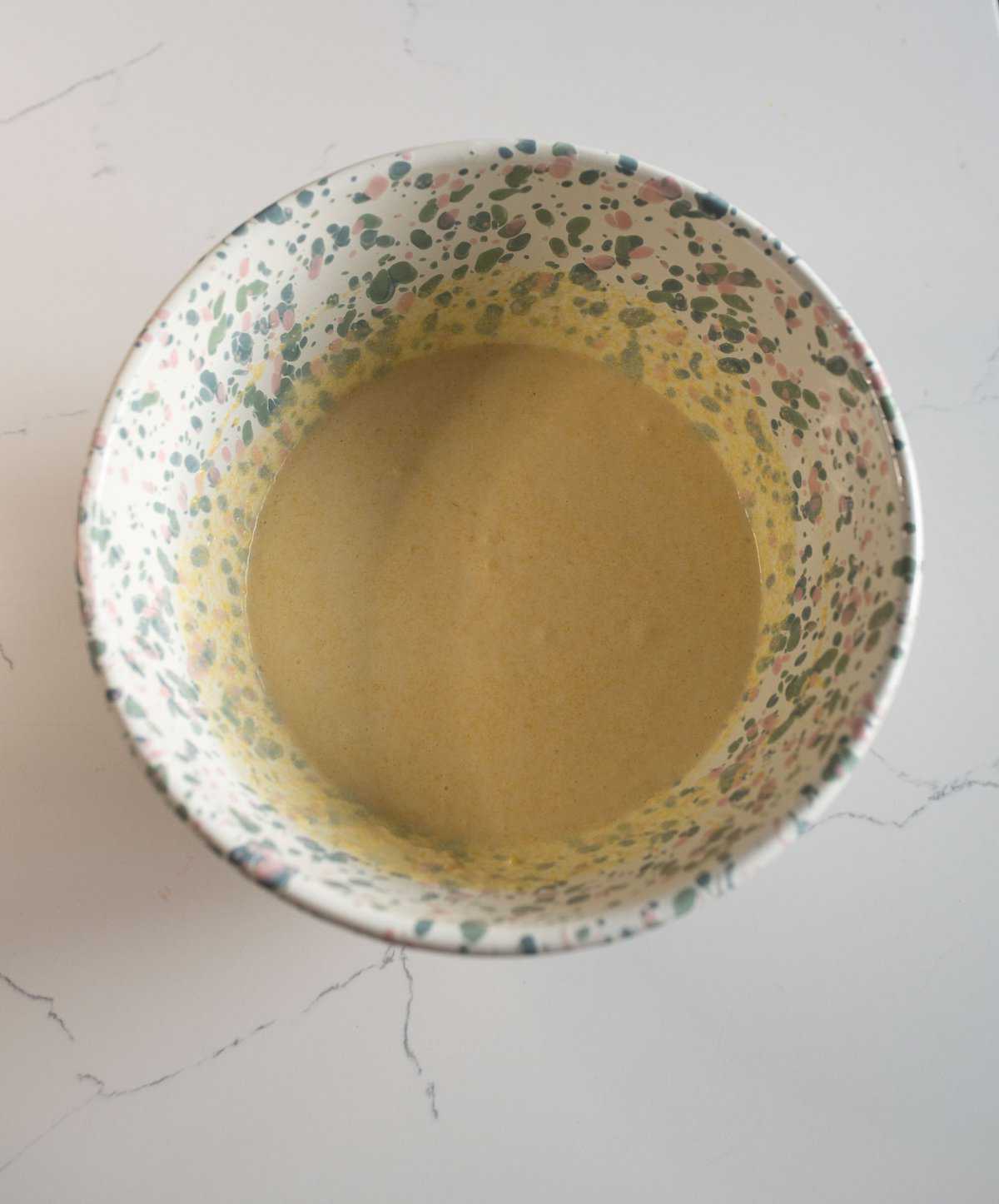 A speckled, colorful bowl on a white marble surface contains a light brown, creamy liquid.