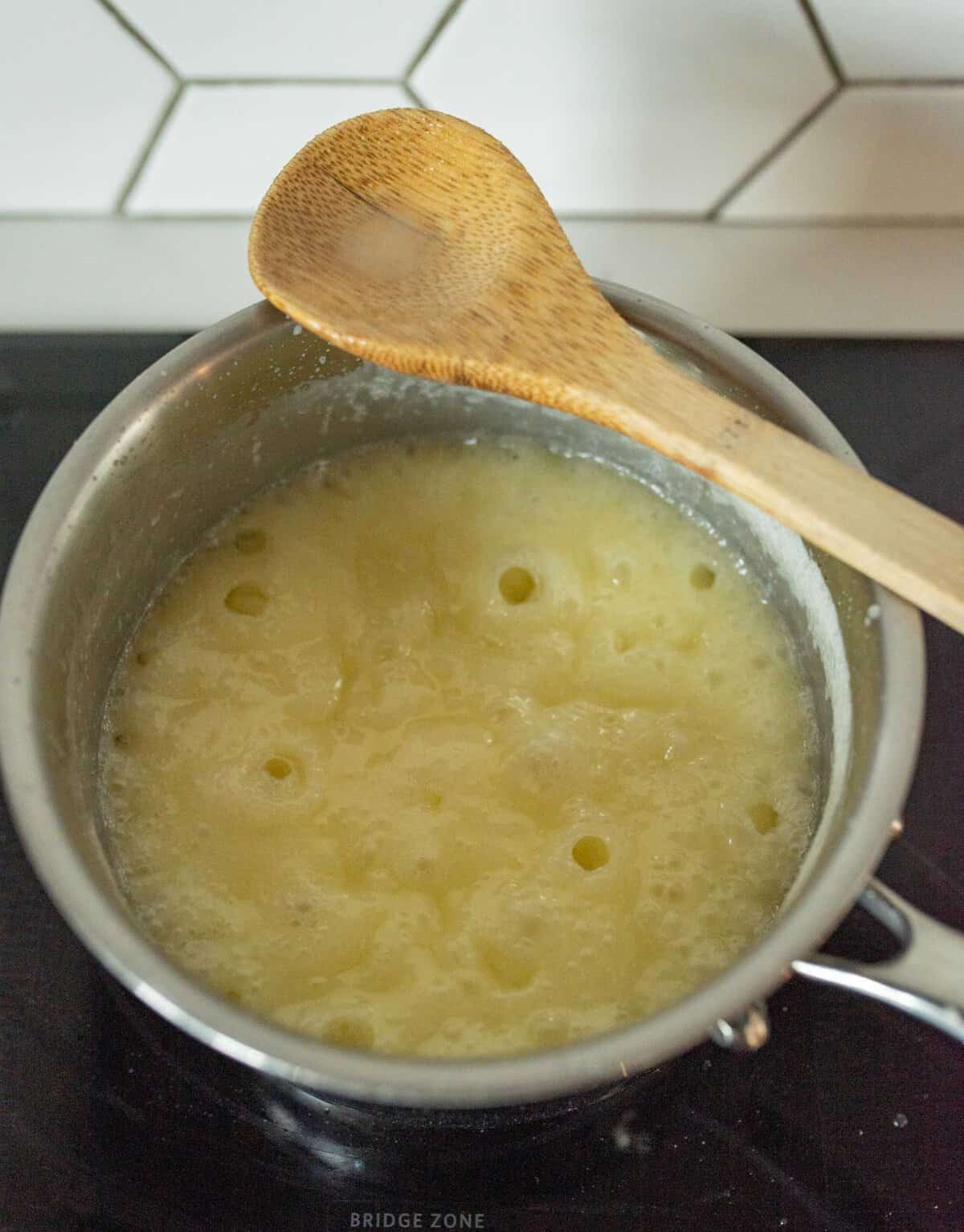 A saucepan on a stove contains bubbling yellow batter. A wooden spoon rests across the top. The backsplash has a white hexagonal tile pattern.