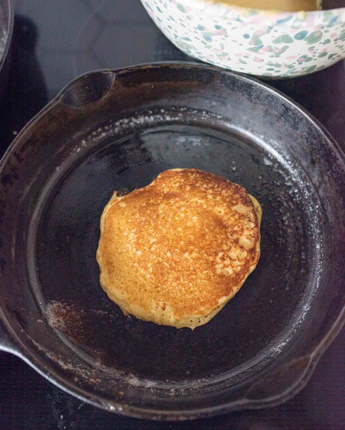 A single golden-brown pancake cooking in a black cast-iron skillet. The pancake appears fluffy with a slightly crisp edge. In the background, part of a colorful bowl is visible, likely containing more batter or pancakes.