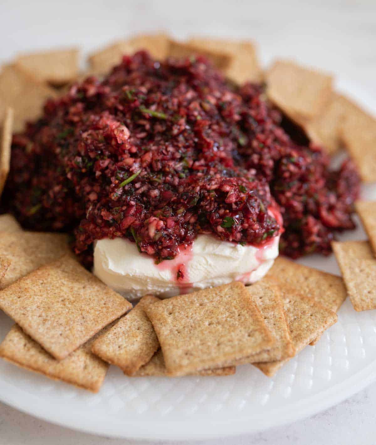 A plate of crackers surrounds a mound of creamy cheese topped with a chunky cranberry relish. The relish is red and finely chopped, spreading slightly over the cheese, creating a vibrant appetizer presentation.