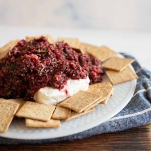 A plate topped with a mound of cranberry salsa on cream cheese, surrounded by square crackers. The setting is on a wooden table with a blue cloth underneath the plate.