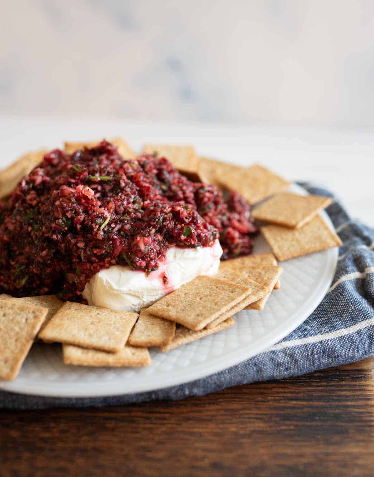 A plate of square crackers surrounds a pile of red and green salsa atop a block of cream cheese. The dish rests on a blue cloth against a light background, with the wooden table partially visible.