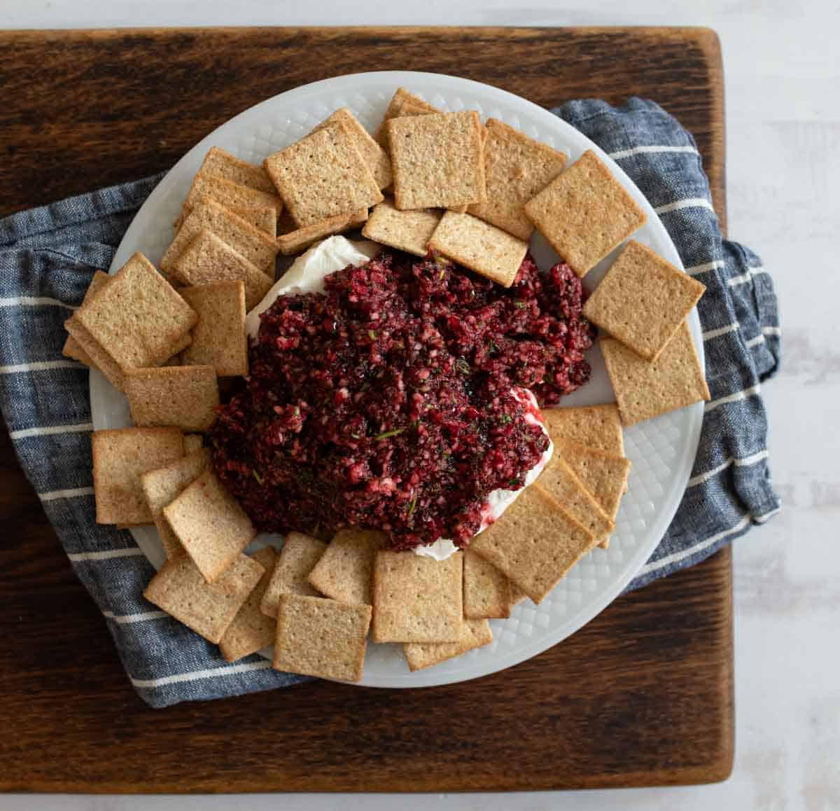 A white plate with a creamy spread topped with a chunky beet mixture, surrounded by square crackers. The plate is on a blue and white cloth on a wooden board.