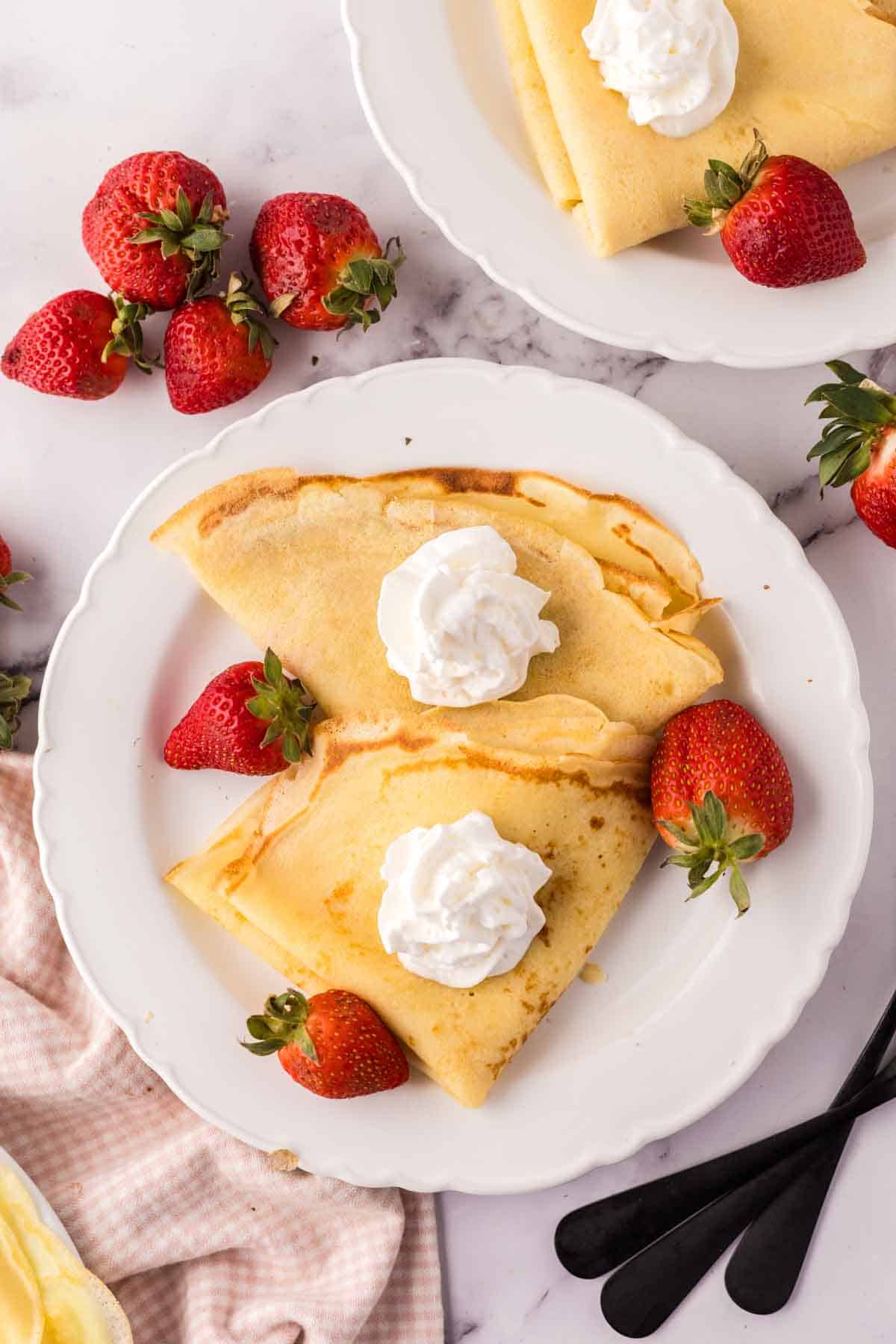 folded crepes on a white plate with fresh berries around and whipped cream.