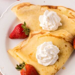 folded crepes on a white plate with fresh berries around and whipped cream.