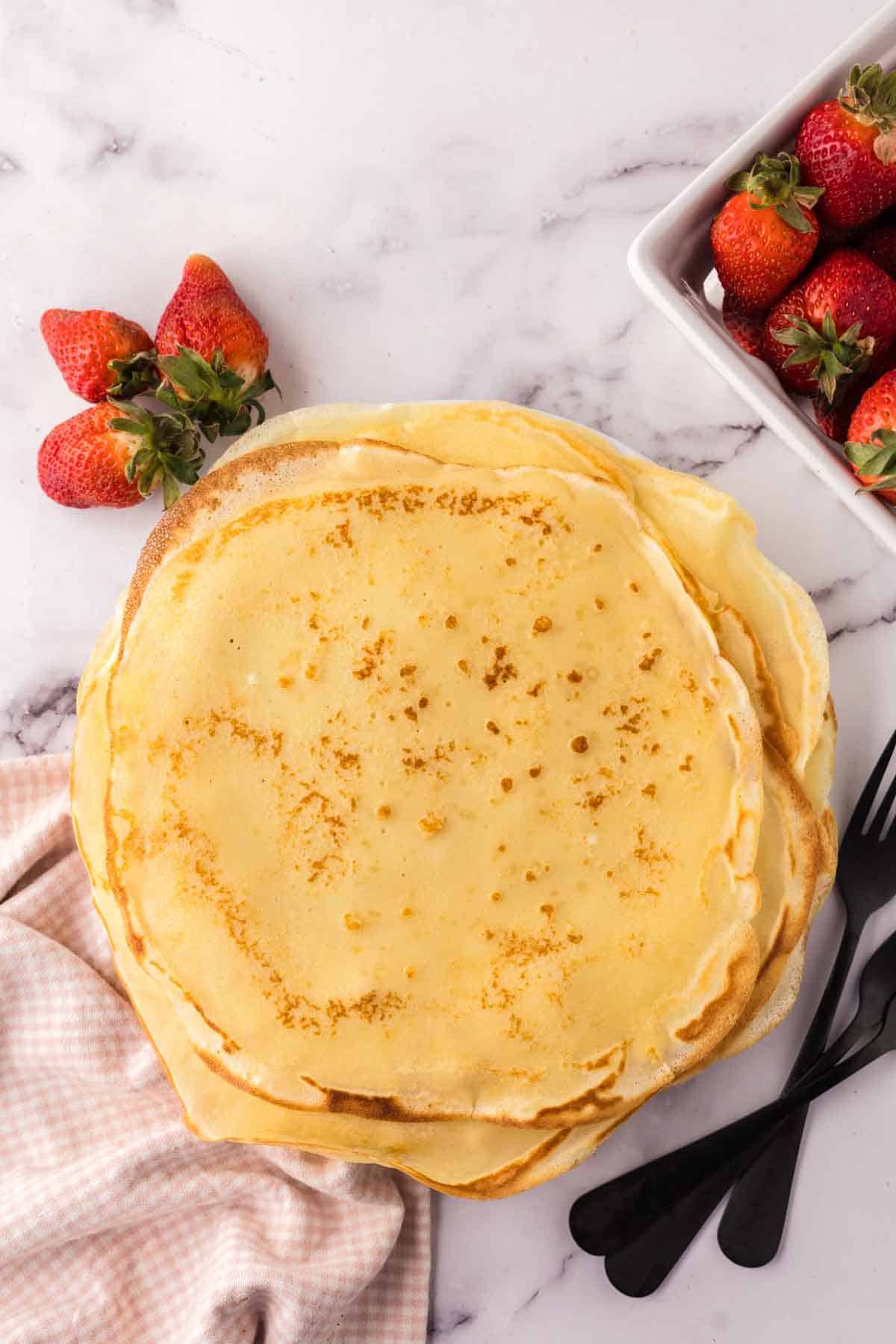 stack of fresh round crepes with strawberries and forks to the side.