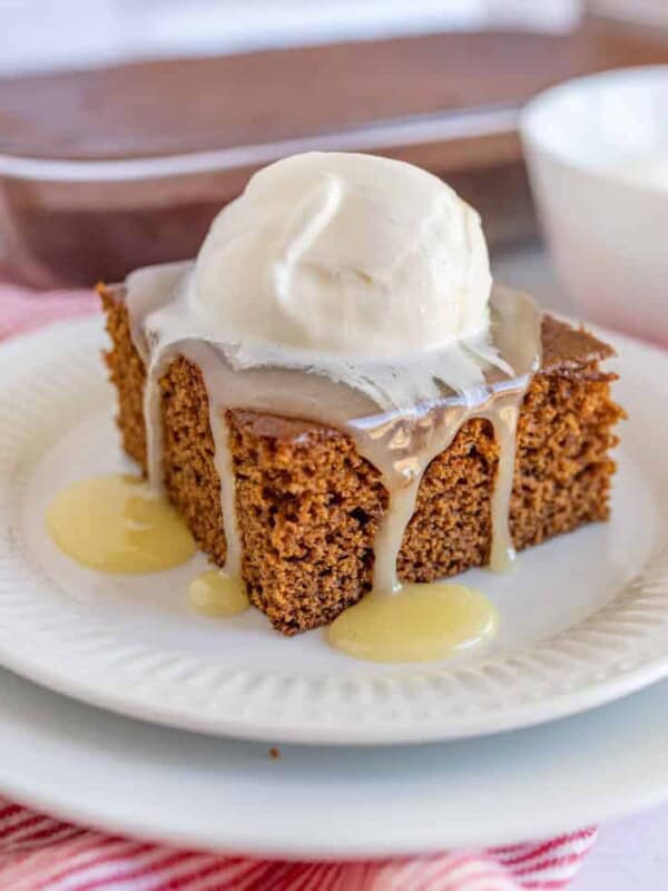 A square piece of gingerbread cake on a white plate topped with vanilla caramel sauce and whipped cream