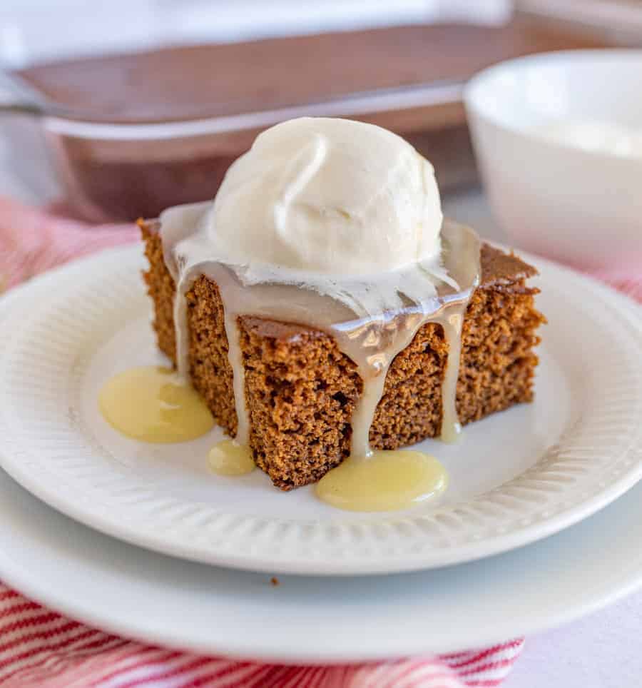 A square piece of gingerbread cake on a white plate topped with vanilla caramel sauce and whipped cream