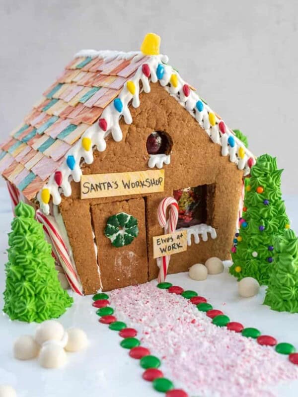 A gingerbread house recipe brings to life Santa's Workshop, featuring colorful candy roof tiles, icing, peppermint poles, and a sign reading "Elves at Work." Green iced trees and a candy path lead to the door.