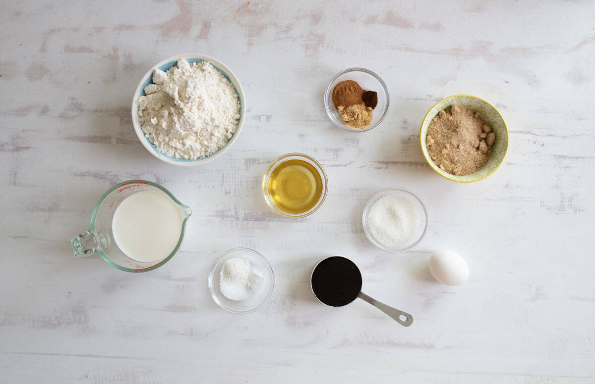 An assortment of baking ingredients arranged on a light surface, including flour, brown sugar, milk, an egg, honey, cinnamon, ginger, sugar, baking soda, salt, and vanilla extract in various bowls and measuring cups.