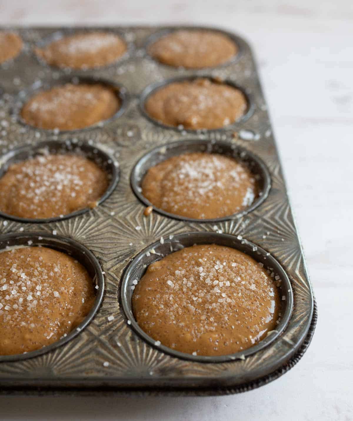 A vintage muffin tin filled with uncooked muffins topped with sugar, ready to be baked. The tin has an ornate pattern and is placed on a lightly textured white surface.