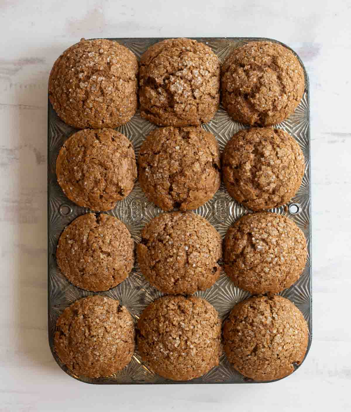 A baking tray filled with twelve freshly baked muffins with a golden-brown, crumbly texture sits on a lightly textured white surface. The muffins are evenly distributed and sprinkled with sugar crystals.