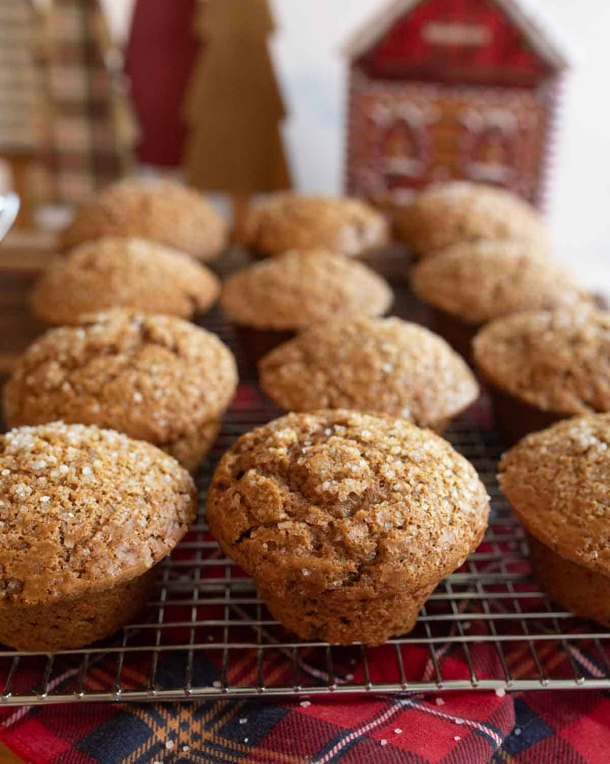 A batch of freshly baked gingerbread muffins topped with sugar crystals resting on a cooling rack. In the background, there are decorative, holiday-themed wooden trees and a red, house-shaped ornament.