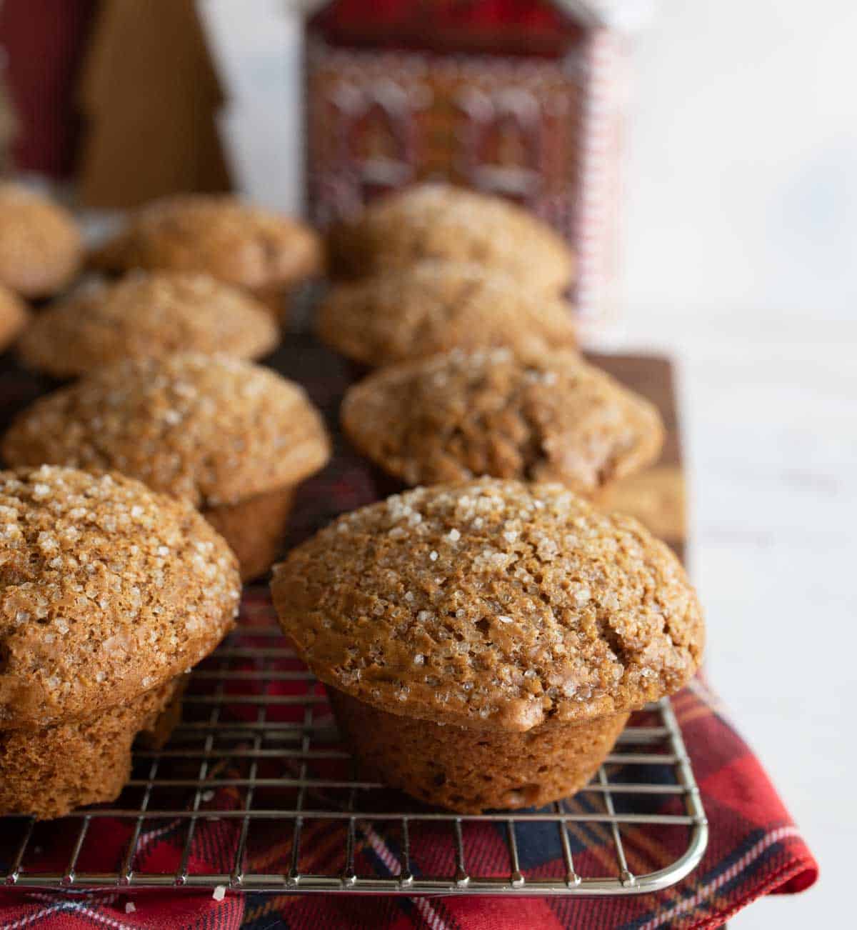 A batch of freshly baked muffins with sugar crystals on top cooling on a wire rack. They are set on a red and black checkered cloth, with a blurred background featuring a decorative tin.