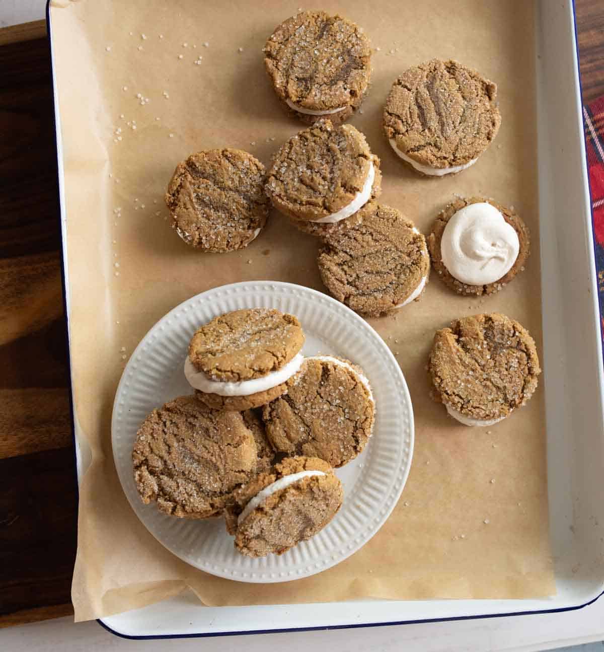 A tray with round sandwich cookies on brown parchment paper. Some cookies are placed on a white plate. They appear to have a creamy filling and a textured brown exterior.