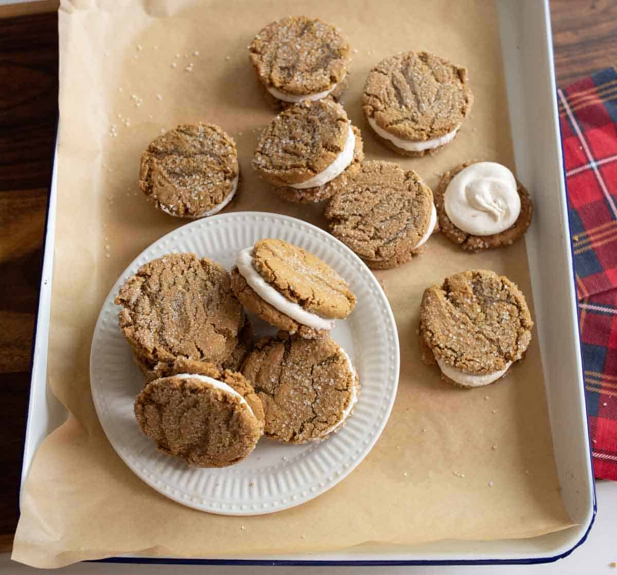 A tray with several ginger molasses sandwich cookies filled with cream, including a white plate holding a stack of these cookies. The tray is lined with parchment paper, and a red plaid cloth is partially visible in the corner.