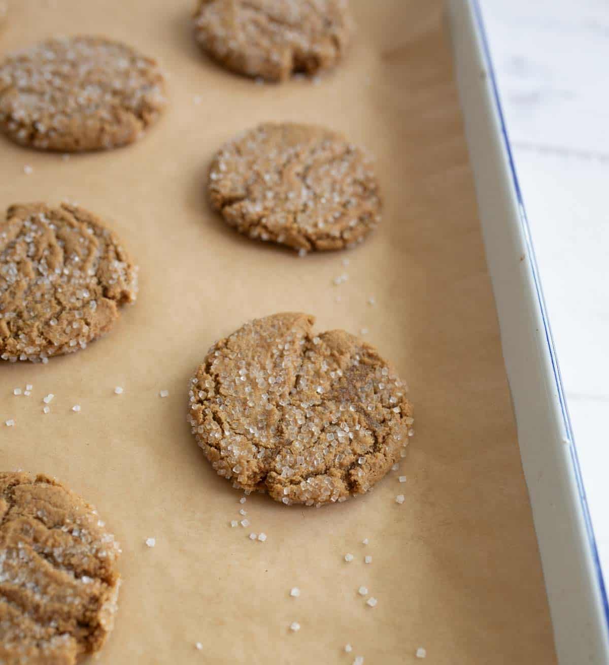 A baking tray lined with parchment paper holds several freshly baked brown cookies sprinkled with coarse sugar. The cookies have a cracked surface and are spaced out evenly. The tray is on a light surface.