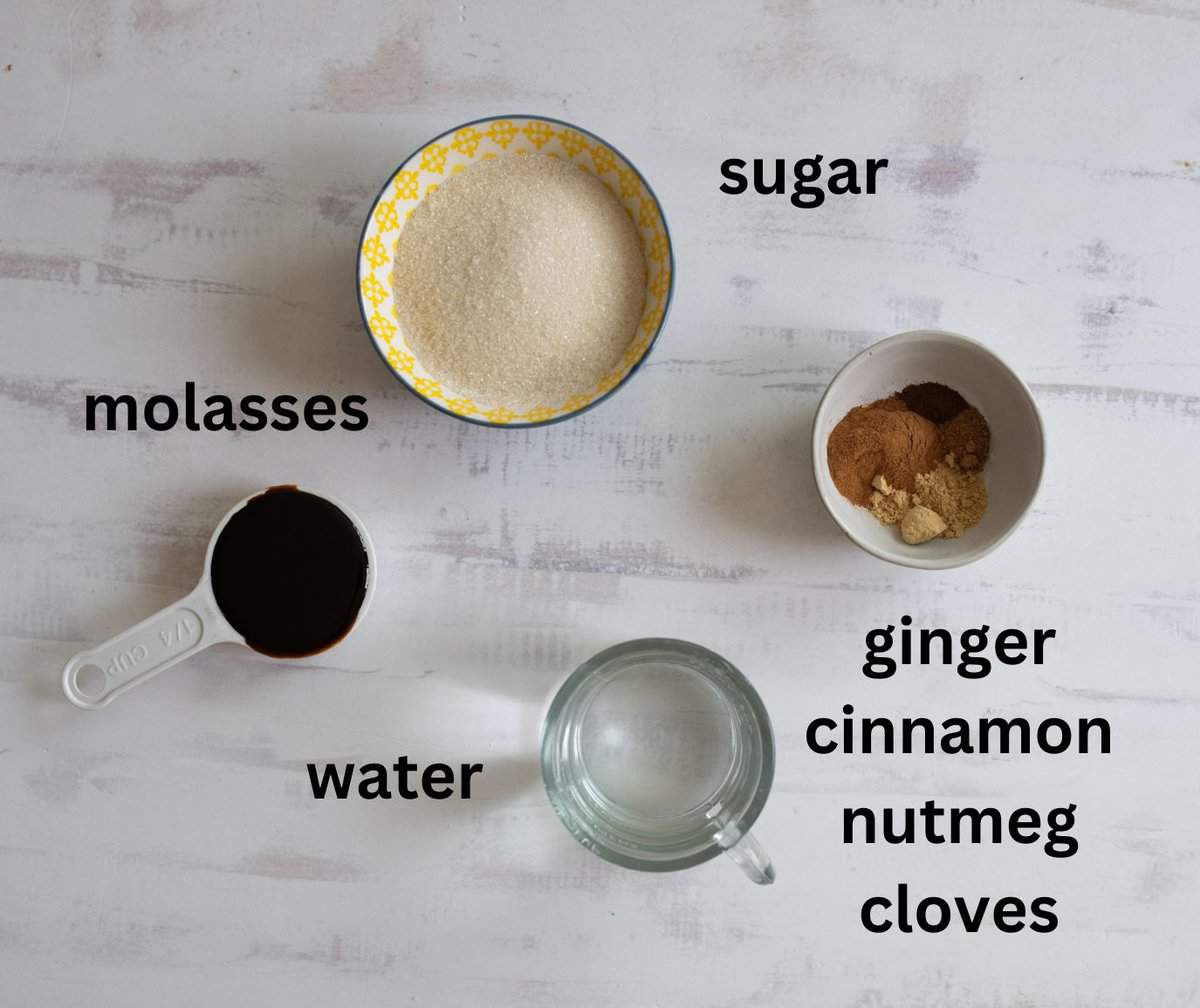 Ingredients for a spice mixture on a white surface: a bowl of sugar, a bowl with ginger, cinnamon, nutmeg, and cloves, a cup of molasses, and a glass of water, all labeled.