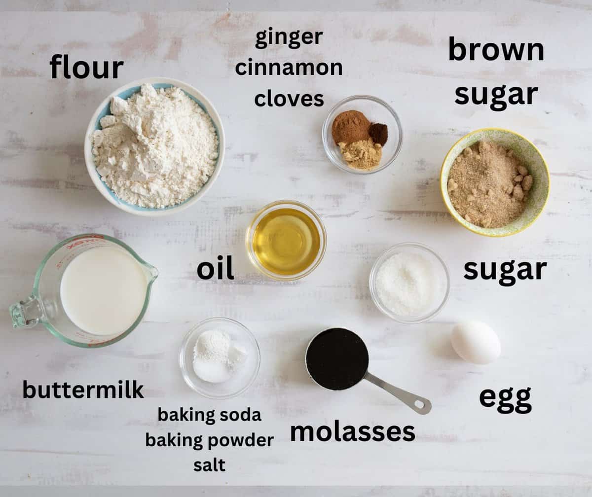 A variety of baking ingredients are displayed on a white wooden surface. These include flour, brown sugar, sugar, oil, buttermilk, molasses, an egg, ginger, cinnamon, cloves, baking soda, baking powder, and salt. Each is labeled.