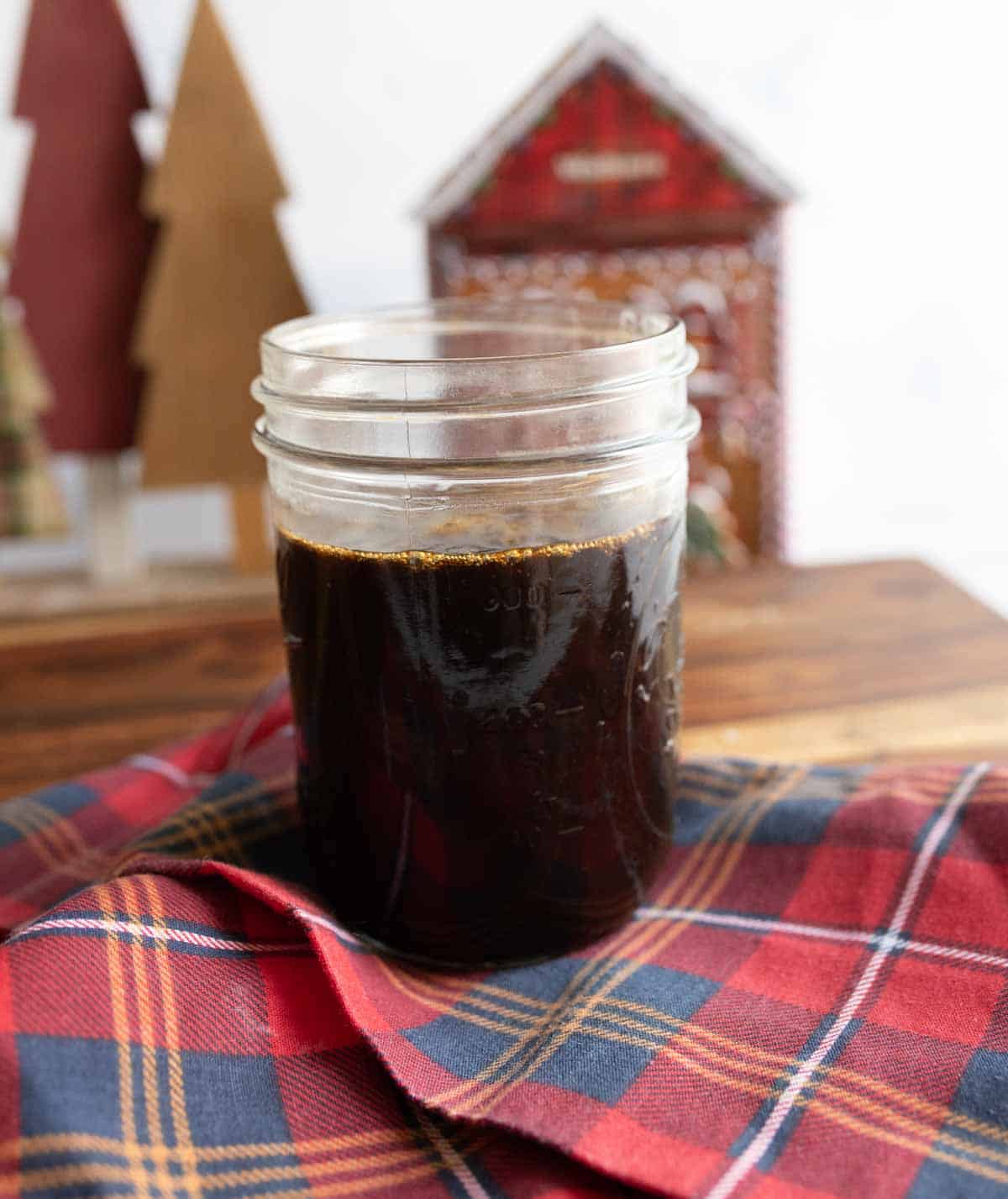 A glass jar filled with dark liquid sits on a red and blue plaid cloth. In the background, there are decorative wooden trees and a small, house-shaped ornament. The setting appears warm and festive.