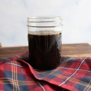 A glass jar filled with dark liquid sits on a folded red and blue plaid cloth. The jar is placed on a wooden surface, and the background is a soft white.