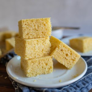 A stack of four square pieces of gluten free cornbread sits on a white plate. The cornbread has a light, fluffy texture and a golden yellow color. A fork and more pieces of cornbread are visible in the blurred background.