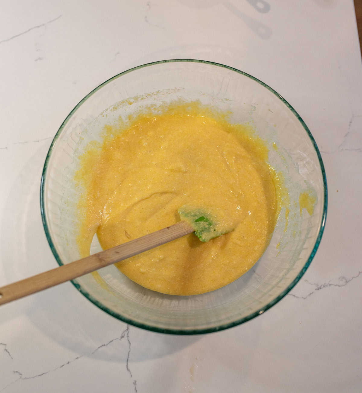 A glass bowl filled with bright yellow cornbread batter, placed on a light marble countertop. A wooden spatula with a green tip rests in the batter.