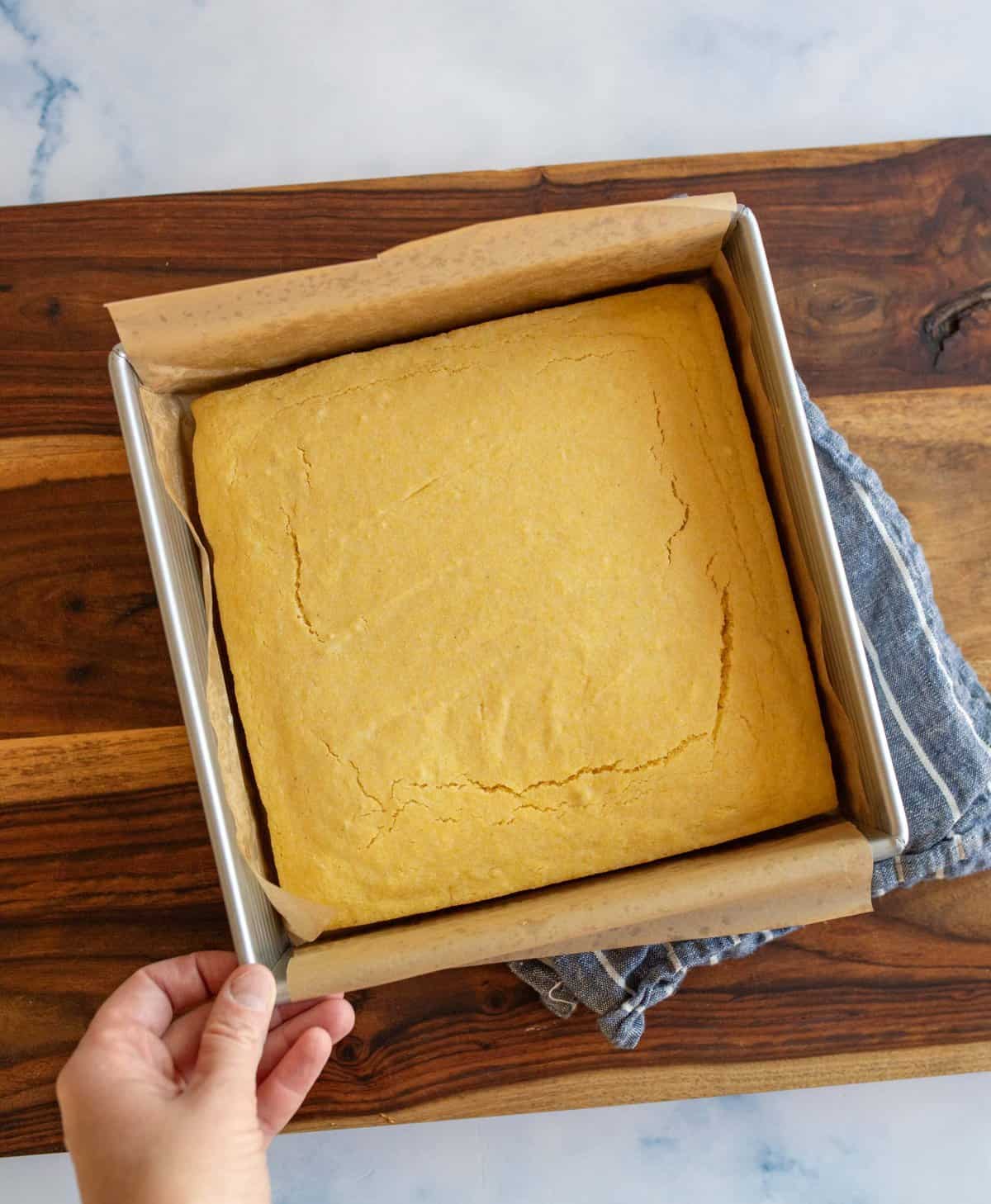 A hand holding a baking pan with freshly baked cornbread resting on a wooden cutting board. The cornbread is a golden-brown square and sits on parchment paper. A blue kitchen towel is partially visible underneath the pan.