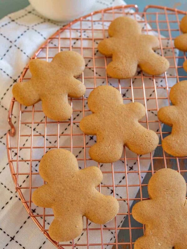 Gluten free gingerbread cookies shaped like people cool on a round copper wire rack. They rest on a white cloth with a black grid pattern. The unadorned cookies are evenly spaced, inviting the warmth of the season into your home.