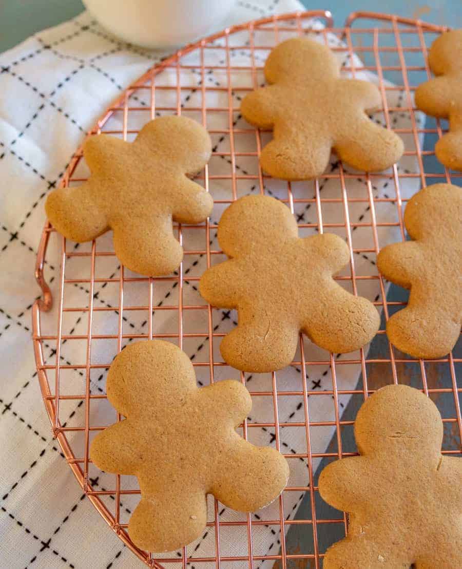 Gluten free gingerbread cookies shaped like people cool on a round copper wire rack. They rest on a white cloth with a black grid pattern. The unadorned cookies are evenly spaced, inviting the warmth of the season into your home.