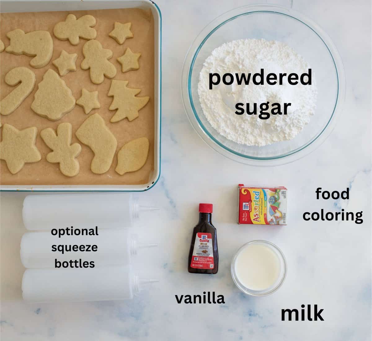 Top view of cookie decorating setup with baked cookies in holiday shapes, a bowl of powdered sugar, milk in a cup, vanilla extract, food coloring, and optional squeeze bottles on a light marble surface.