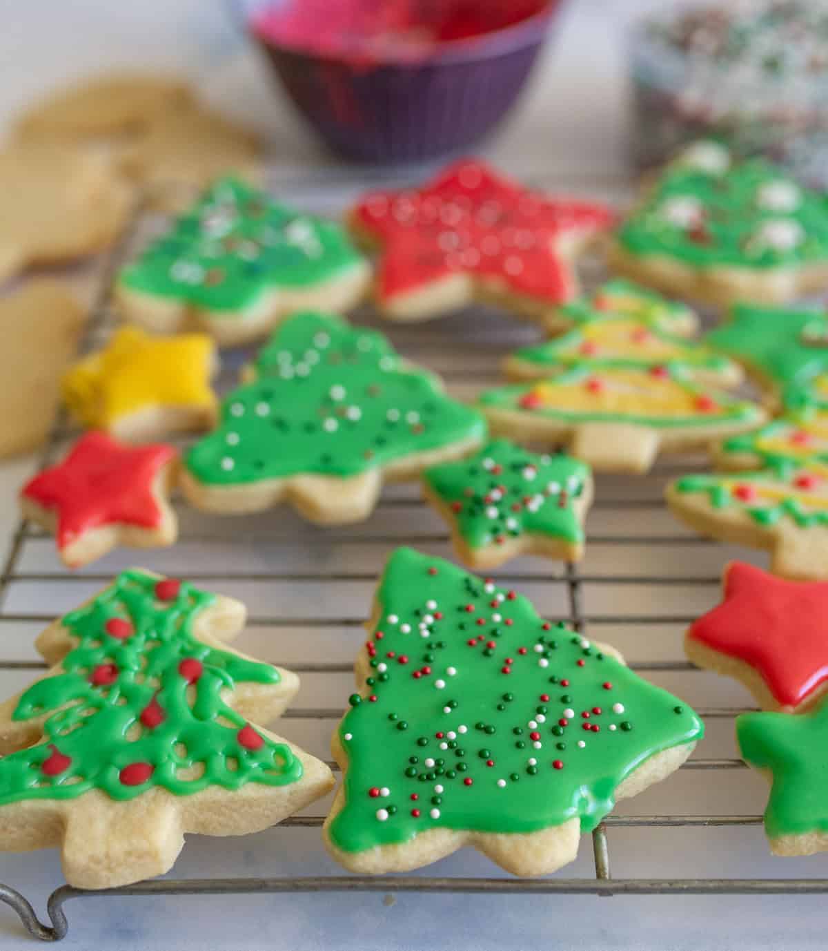 A rack of holiday cookies shaped like trees and stars, decorated with colorful icing and sprinkles. Theres a variety of green, red, and yellow icing on the cookies, with bowls of sprinkles in the background.