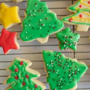 Assorted festive sugar cookies in the shapes of Christmas trees and stars, decorated with a sugar cookie icing recipe in green, red, and yellow hues, plus colorful sprinkles, resting on a cooling rack.