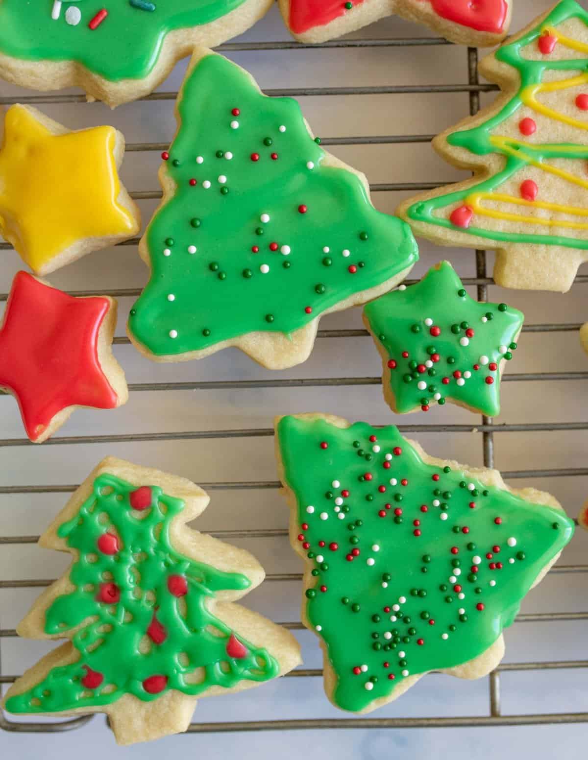 Christmas-themed cookies in various festive shapes, including Christmas trees and stars, are decorated with green, red, and yellow icing and sprinkles, and rest on a cooling rack.