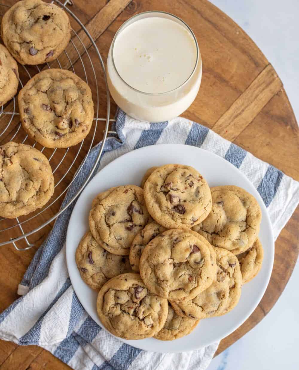 top view of a plate stacked with chocolate chip cookies with milk and some other cookies on a drying rack.