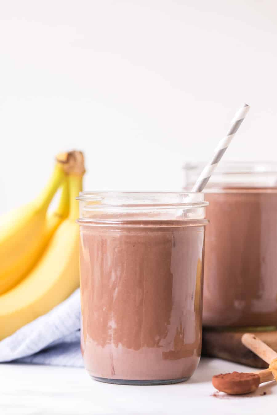 two glass canning jars filled with peanut butter chocolate smoothies with paper straws next to two bananas and teaspoon of cinnamon