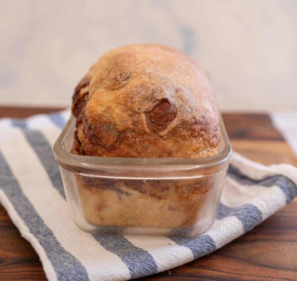 close up shot of baked sourdough cinnamon swirl bread in clear bread pan on blue and white striped towel.