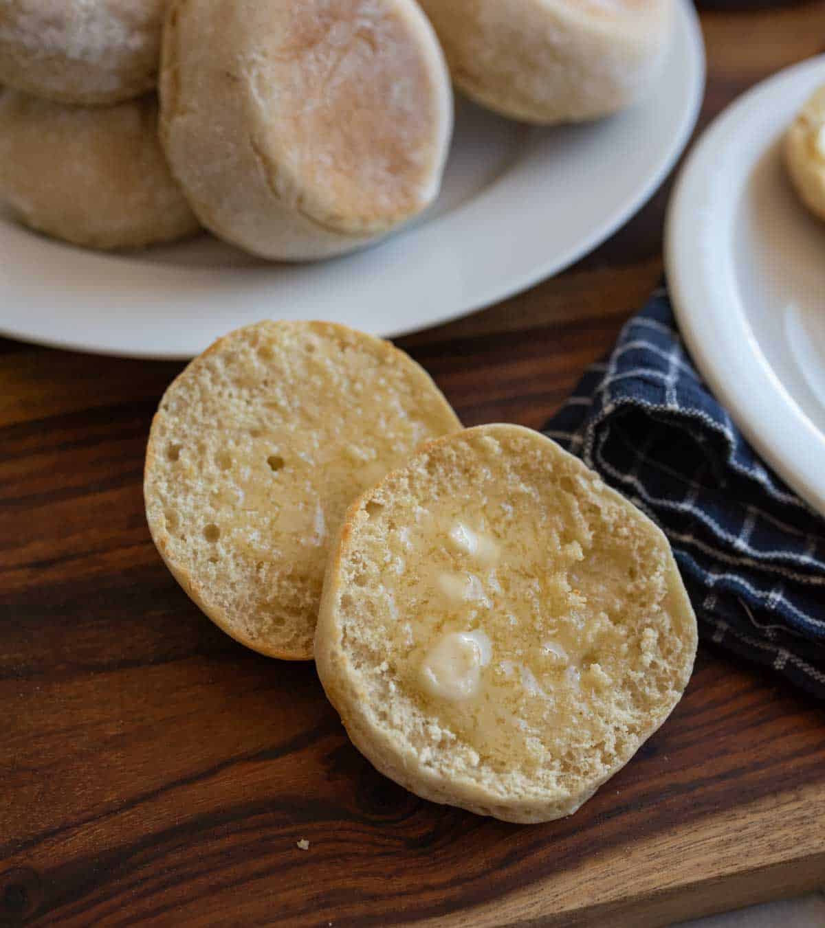 baked english muffin split with melting butter on it and pile of muffins on white plate in background.