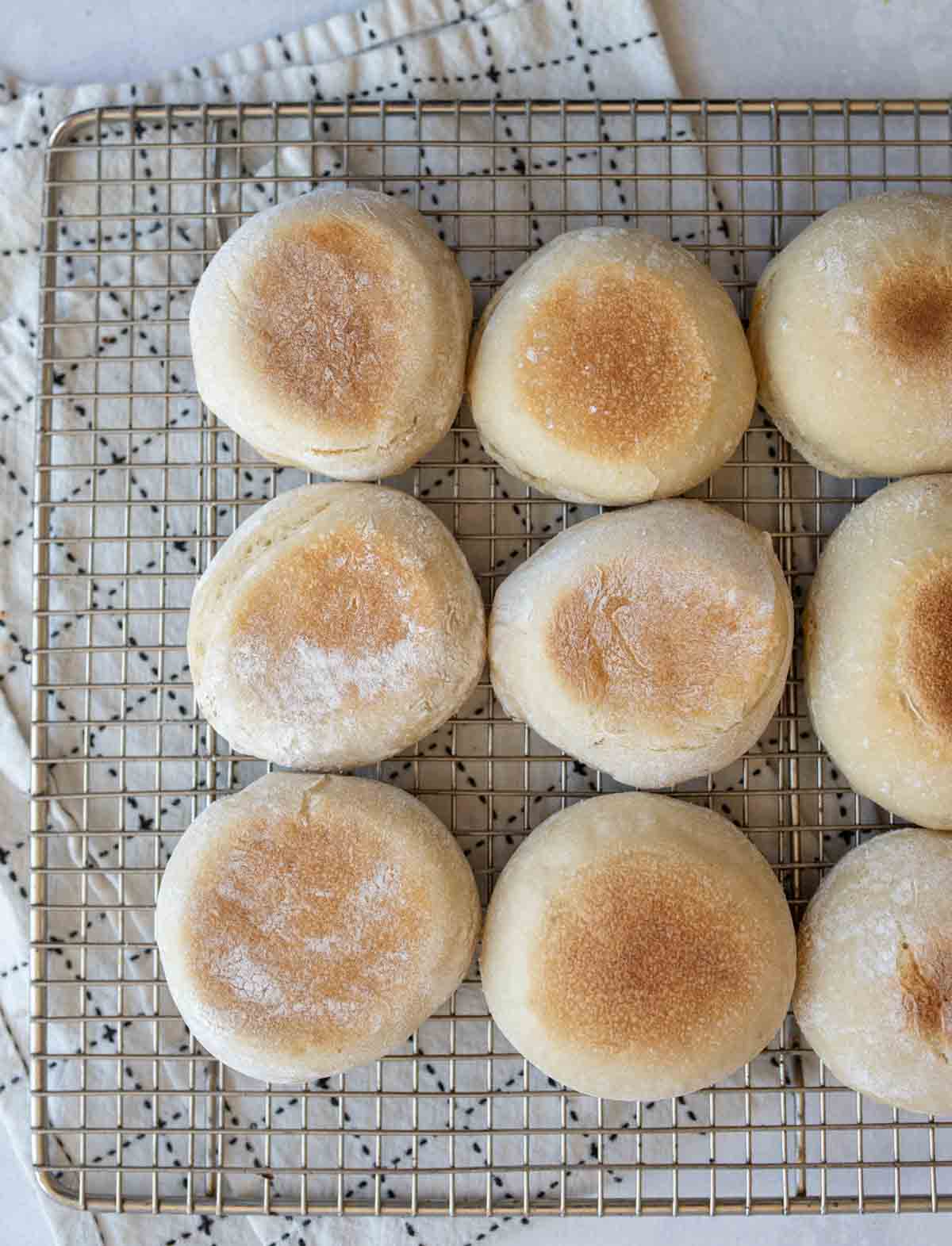 baked sourdough english muffins on metal wire cooling rack on black and white towel.